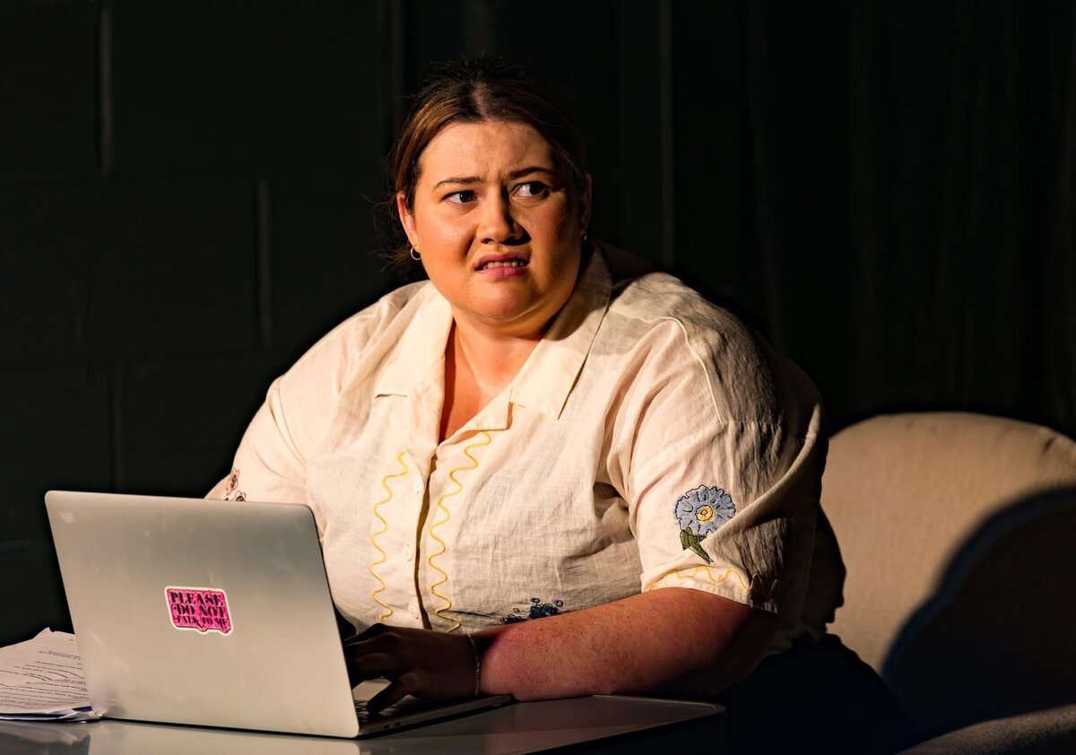 Sarah McNally in a theatre production, seated at a laptop